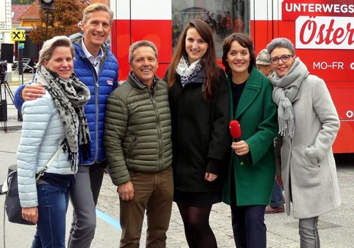 Eine Gruppe von fünf Erwachsenen, darunter zwei Frauen mit Mikrofonen, steht vor einem roten Zug, lächelt und posiert für ein Foto. Im Hintergrund sind Bäume und ein Gebäude mit dem Wort 'UNIT' zu sehen.