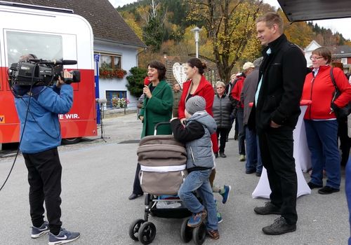 Eine Frau hält ein Mikrofon und spricht bei einer Veranstaltung im Freien. Ein Junge in einer Jacke schiebt einen Kinderwagen. Menschen versammeln sich, einige tragen Mützen. Bäume und ein Gebäude sind im Hintergrund.