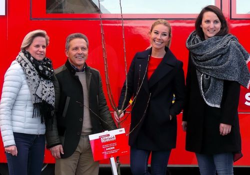 Vier Personen stehen neben einem roten Bus und lächeln für ein Foto. Ein Mann hält einen jungen Baum in der Hand, neben ihm eine Frau. Eine Frau mit Schal lächelt rechts in die Kamera.
