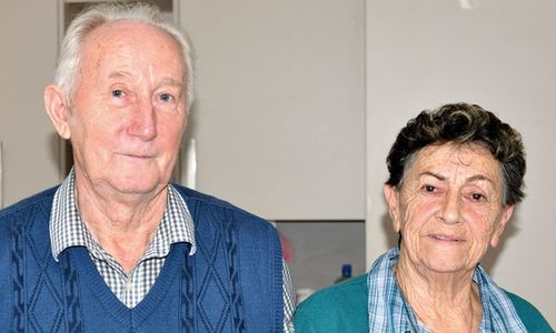 Zwei ältere Menschen lächeln, ein Mann mit weißen Haaren und eine Frau mit lockigen Haaren, stehen eng beieinander. Sie sind in einer Küche.