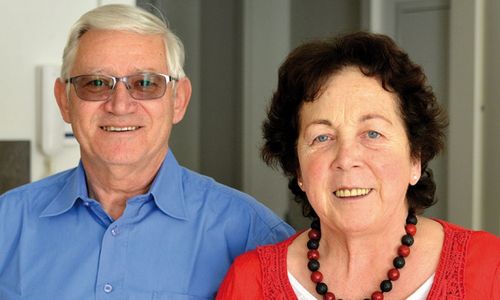 Ein älterer Mann und eine ältere Frau lächeln in die Kamera und stehen eng beieinander. Der Mann trägt eine Brille und ein blaues Hemd, die Frau ein rotes Oberteil und eine rote und schwarze Perlenkette.