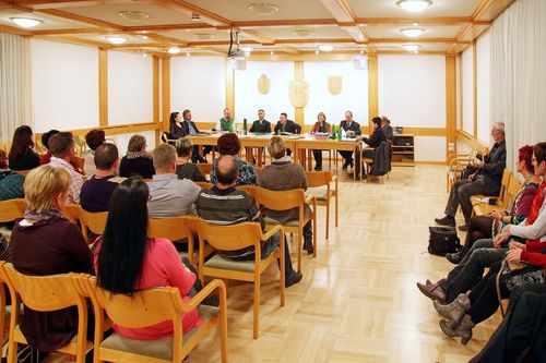Eine Konferenz mit einem Podium mit sechs Personen, die an einem Tisch sitzen und ein Publikum von etwa 15 Personen in einem Raum mit Holzwänden und -böden ansprechen.