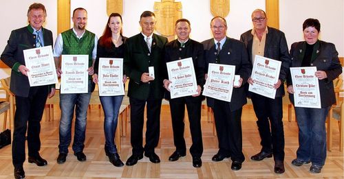 Sechs Menschen stehen in einer Reihe, feierlich gekleidet, lächelnd und halten Urkunden. Der Hintergrund zeigt einen Innenraum mit Holzboden und einer dekorativen Wand.