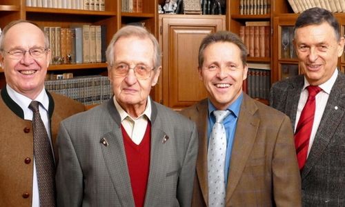 Vier Männer stehen in einer Bibliothek und lächeln. Der Mann auf der linken Seite trägt eine Brille, ein rotes Hemd und einen grauen Anzug. Der Mann in der Mitte trägt ein blaues Hemd und einen braunen Anzug. Der Mann auf der rechten Seite trägt einen braunen Anzug und eine Krawatte.