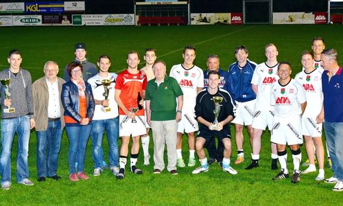 Eine Fußballmannschaft in weißen und roten Trikots posiert mit Trophäen auf einem Feld, mit zwei Männern in grünen und blauen Jacken in der Mitte, und Zuschauern dahinter.