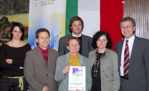 Eine Gruppe von Menschen in Anzügen und Krawatten steht vor einem Hintergrund mit italienischer Flagge. Eine Frau hält ein Zertifikat, und ein Mann lächelt in der Mitte.