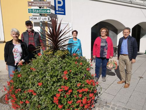 Fünf Personen, darunter drei Frauen und zwei Männer, stehen vor einem Gebäude mit Straßenschildern, die auf ein Museum, ein Stadion und ein Labor hinweisen.