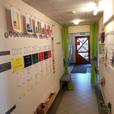 Ein Flur mit Bulletin Boards, einer Bank und einem Teppich vor einer Tür. Eine Lampe ist an der Decke und Bücher sind auf den Regalen.