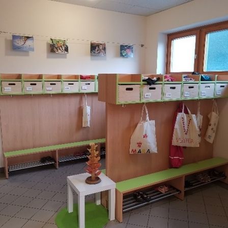 Ein Raum mit Holzschränken, Schuhen und Taschen. Ein weißer Tisch und ein grüner Teppich liegen auf dem Boden. Fenster sind an der Wand.