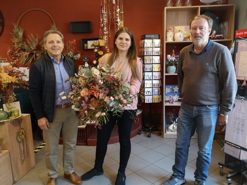 Drei Personen stehen in einem Raum, wobei eine Frau einen Blumenstrauß hält. Der Mann auf der linken Seite trägt eine Jacke und Hose. Der Mann auf der rechten Seite trägt ein Hemd und Jeans. Dahinter befindet sich ein Regal mit verschiedenen Gegenständen.