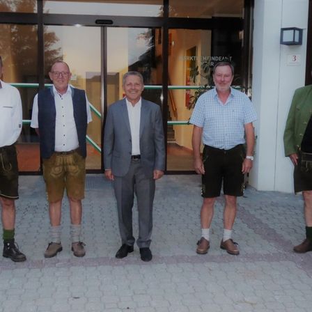 Fünf Männer stehen vor einem Gebäude. Sie tragen alle Schuhe. Der Mann in der Mitte trägt einen grauen Anzug. Die anderen sind in traditioneller Kleidung gekleidet.