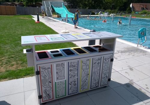 Ein Mülleimer mit mehreren Fächern steht in der Nähe eines Schwimmbeckens. Menschen schwimmen im Becken, und eine Rutsche ist in der Nähe. Der Mülleimer hat Illustrationen von recycelbaren Gegenständen.