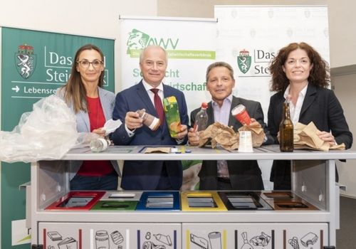 Vier Personen in formeller Kleidung stehen hinter einem Recyclingbehälter und halten jeweils verschiedene Gegenstände wie Plastik- und Papiertüten in der Hand, mit Bannern im Hintergrund, die 'Das Steirische Wirtschaftsverband' und 'Das Stei' lesen.