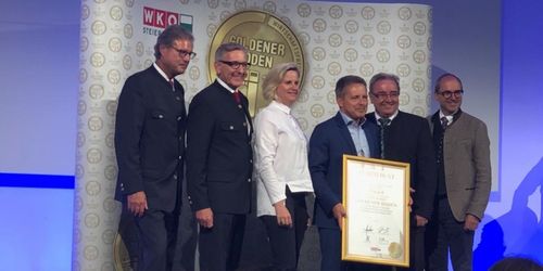 Fünf Personen stehen zusammen und halten eine goldene Plakette. Sie scheinen eine Auszeichnung zu erhalten. Der Mann ganz rechts hält die Plakette, während die anderen lächeln. Hinter ihnen befindet sich eine weiße Wand mit vielen goldenen Logos.