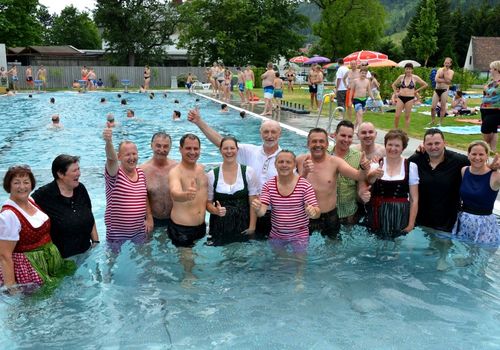 Eine Gruppe von Erwachsenen, sowohl Männer als auch Frauen, posiert im Pool mit erhobenem Daumen. Einige tragen Badeanzüge, andere traditionelle Kleidung. Der Hintergrund zeigt einen überfüllten Pool, Grünflächen und Sonnenschirme.