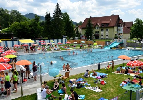 Ein Freibad mit Schwimmenden und Sonnenbadenden. Sonnenschirme und Liegestühle sind auf der Wiese aufgestellt. Im Hintergrund befinden sich Häuser und Bäume.
