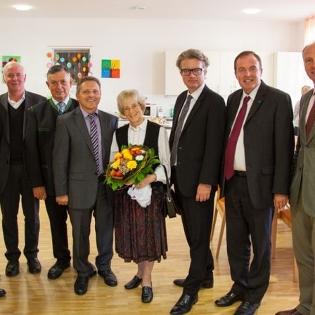 Eine Gruppe von Menschen steht in einem Raum und posiert für ein Foto. Eine Frau hält einen Blumenstrauß.