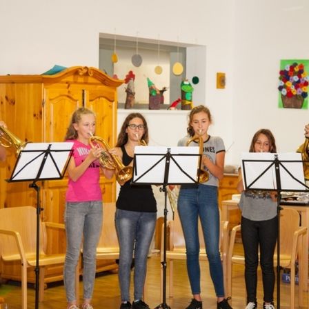 Eine Gruppe junger Kinder und zwei erwachsene Männer spielen Trompeten in einem Klassenzimmer mit Musiknoten auf Ständern vor ihnen.