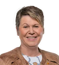 Eine Frau mit kurzen Haaren und einer braunen Jacke lächelt in die Kamera. Sie trägt ein weißes Shirt mit blauem Design. Sie steht vor schwarzem Hintergrund.
