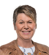 Eine Frau mit kurzen Haaren und einer braunen Jacke lächelt in die Kamera. Sie trägt ein weißes Shirt mit blauem Design. Sie steht vor schwarzem Hintergrund.
