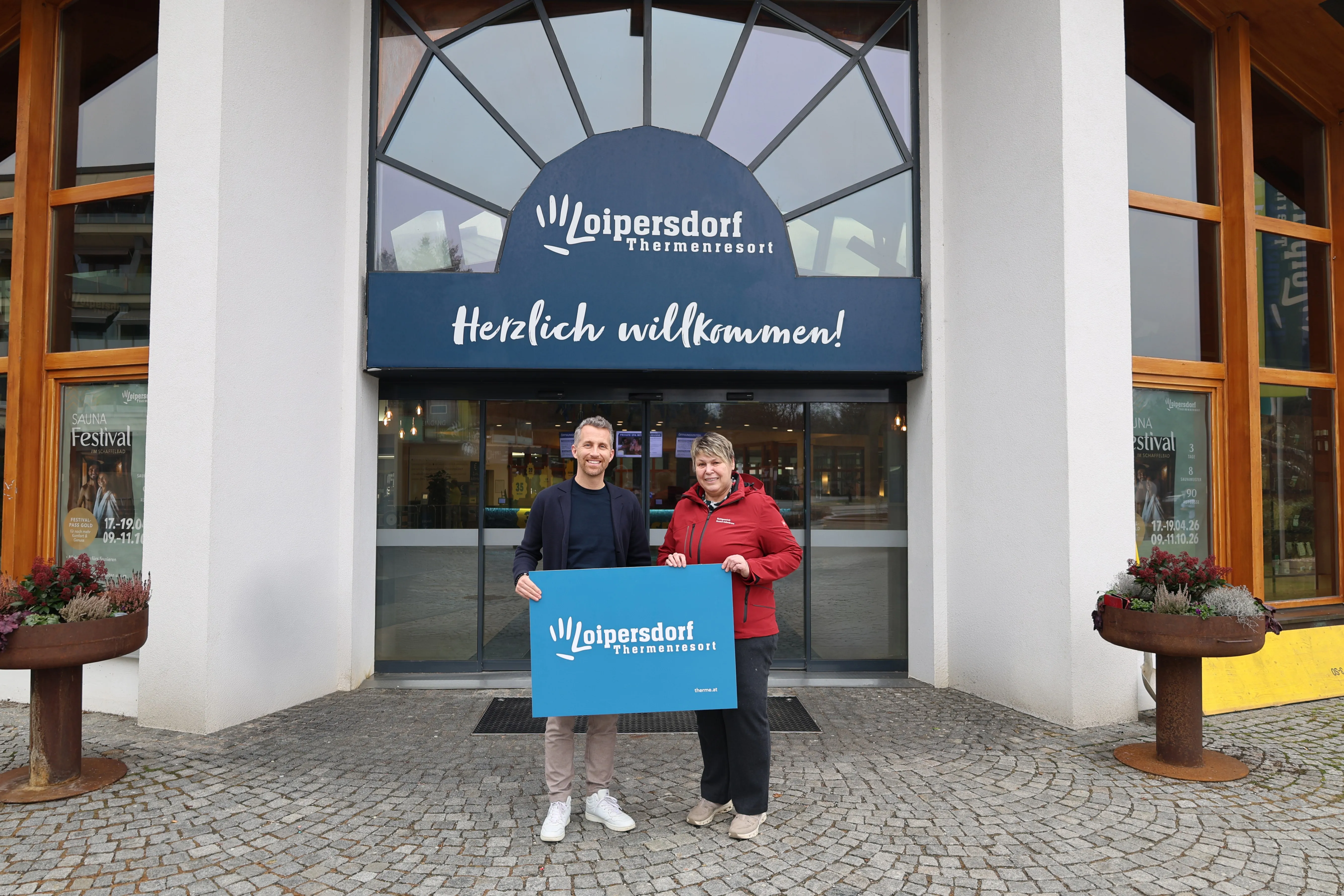 Ein Mann und eine Frau stehen vor einem Gebäude und halten ein Schild. Das Gebäude ist ein Loipersdorf Thermal Resort. Sie lächeln.