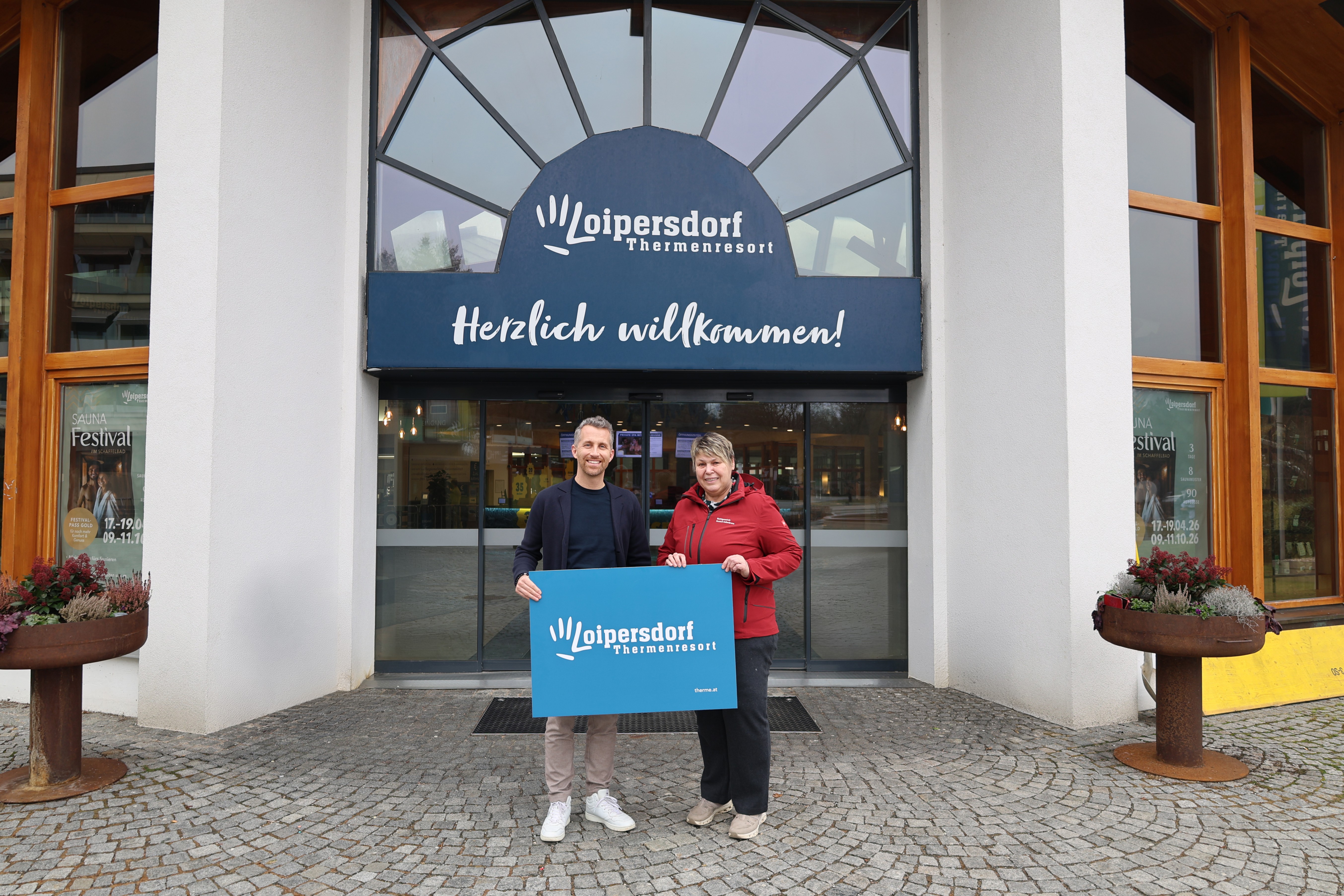 Ein Mann und eine Frau stehen vor einem Gebäude und halten ein Schild. Das Gebäude ist ein Loipersdorf Thermal Resort. Sie lächeln.