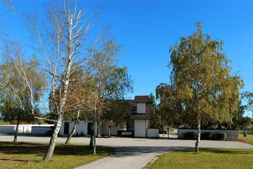 Ein Haus mit braunem Dach steht auf einer Rasenfläche. Bäume mit kahlen Ästen umgeben es. Eine Einfahrt führt zum Eingang. Der Himmel ist klar und blau.