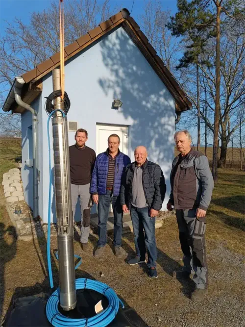 Vier Männer stehen vor einem Haus. Einer hat eine Jacke mit einem gestreiften Muster. Sie lächeln.