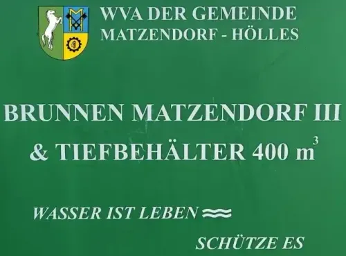 Ein grünes Schild mit dem Text 'WWA der Gemeinde Matzendorf-Hölles' und einem Logo. Darunter steht 'Brunnen Matzendorf III & Tiefbehälter 400 m' und 'Wasser ist Leben'. Unten steht 'Schütze es'.