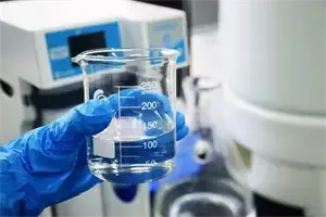 Eine Person in blauen Handschuhen hält ein Becherglas mit einer Flüssigkeit, wahrscheinlich führt sie ein Experiment in einem Labor mit Maschinen und Geräten im Hintergrund durch.