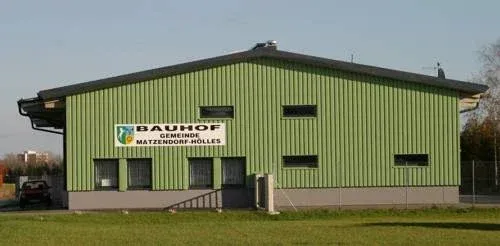 Ein grünes Gebäude mit einem Schild, auf dem 'Bauhof Gemeinde Matzendorf-Hölles' steht, hat ein geneigtes Dach und einen eingezäunten Bereich. Es hat einige Fenster und ein Auto, das draußen geparkt ist.