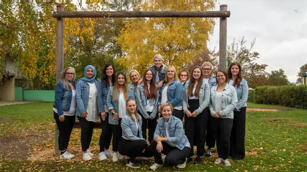 Eine Gruppe von Frauen in Jeansjacken und Turnschuhen posiert für ein Foto in einem Park mit Bäumen und einer Holzstruktur im Hintergrund.