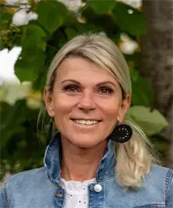 Eine Frau mit blondem Haar lächelt in die Kamera, während sie eine Jeansjacke und schwarze Ohrringe trägt. Sie ist draußen mit einem Baum und grünen Blättern im Hintergrund.