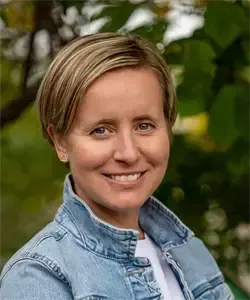 Eine lächelnde Frau mit kurzen blonden Haaren in einer Jeansjacke und einem weißen T-Shirt posiert im Freien.