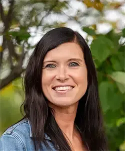 Eine lächelnde Frau mit langen schwarzen Haaren und blauen Augen, die eine Jeansjacke trägt, steht im Freien mit Bäumen im Hintergrund.