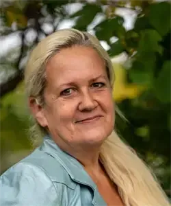 Eine Frau mit blonden Haaren lächelt und posiert für ein Foto vor einem grünen Blatthintergrund. Sie trägt ein hellblaues Hemd.