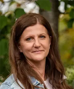Eine Frau mit langen braunen Haaren lächelt in die Kamera und trägt eine Jeansjacke. Sie steht vor einem unscharfen Hintergrund mit Bäumen und Pflanzen.