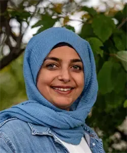 Eine lächelnde Frau mit einem blauen Kopftuch und einer Jeansjacke posiert vor grünen Bäumen.