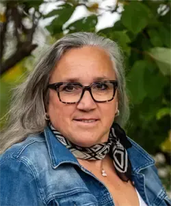 Eine Frau mit grauen Haaren, die eine Brille und eine Jeansjacke mit einem gemusterten Schal trägt, steht vor einem grünen Baum.
