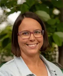 Eine lächelnde Frau mit kurzen Haaren und einer Brille steht draußen und posiert für ein Foto. Sie trägt ein hellblaues Shirt und hat einen Baum und Grünpflanzen im Hintergrund.