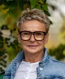 Eine Frau mit kurzen Haaren und einer Brille steht in einem Garten. Sie trägt eine Jeansjacke und ein weißes Hemd. Hinter ihr sind Bäume und Grün verschwommen.