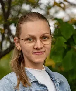 Eine Frau mit blonden Haaren, die eine Jeansjacke und eine Brille trägt, lächelt im Freien mit grünem Hintergrund.