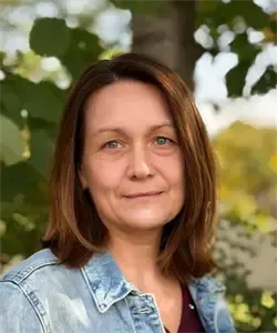 Eine Frau mit kurzen braunen Haaren lächelt für ein Foto vor einem verschwommenen grünen Hintergrund. Sie trägt eine blaue Jeansjacke und ein weinrotes Oberteil.