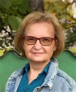 Eine Frau mit kurzen blonden Haaren und einer Brille lächelt für ein Foto. Sie trägt eine Jeansjacke über einem blauen Hemd. Hinter ihr steht eine grüne Bank und Bäume.