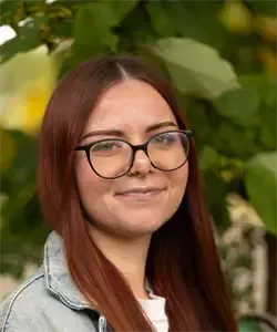 Eine Frau mit langen roten Haaren, Brille und Jeansjacke lächelt für ein Foto, mit grünen Blättern im Hintergrund.