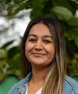 Eine Frau mit langen blonden Haaren lächelt und steht vor einem Baum. Sie trägt eine Jeansjacke und ein weißes Hemd.