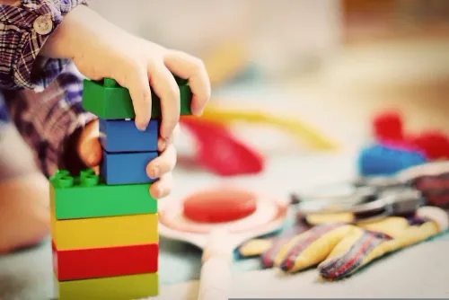 Ein Kind spielt mit einem Stapel bunter Lego-Steine und hält sie mit beiden Händen. Der Tisch ist mit verschiedenen Spielzeugen übersät, darunter Handschuhe.