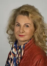 Ein Porträt einer Frau mit blonden, lockigen Haaren, die eine rote Lederjacke und einen gemusterten Schal mit blauen, weißen und orangefarbenen Farben trägt. Sie steht vor einem schlichten weißen Hintergrund.