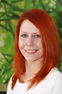 Eine Frau mit roten Haaren und einem weißen T-Shirt lächelt in die Kamera. Sie hat einen Nasenring und steht vor einem grünen Hintergrund.