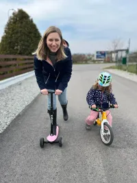 Eine Frau mit lächelndem Gesicht fährt einen rosa Roller, und ein kleines Mädchen fährt ein gelbes Fahrrad auf der Straße. Dahinter befindet sich ein Rasen.